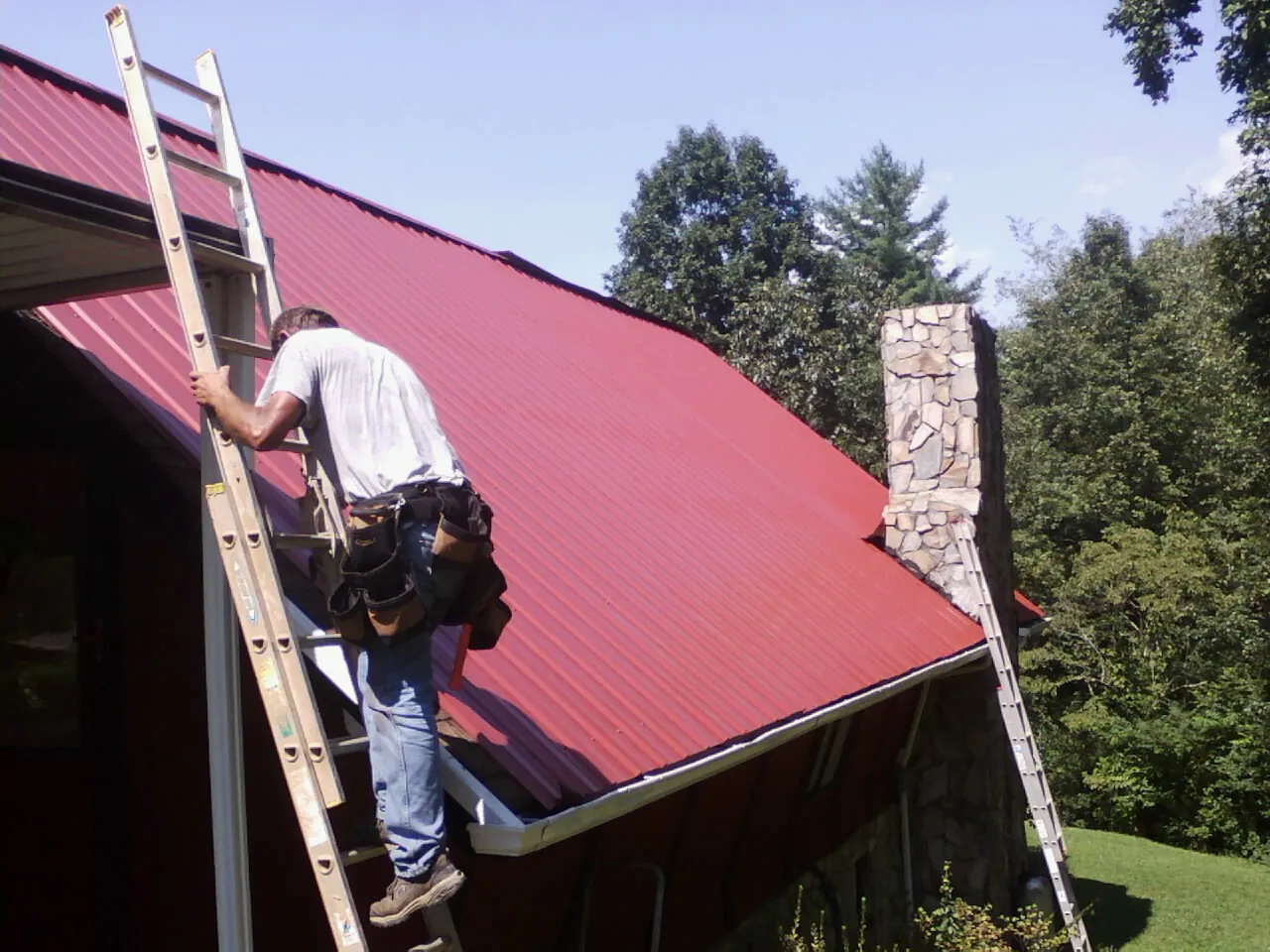 Roofer on a ladder beside a red metal roof and stone chimney — Soffit & Fascia Repair by Roofing Brothers in Nehawka